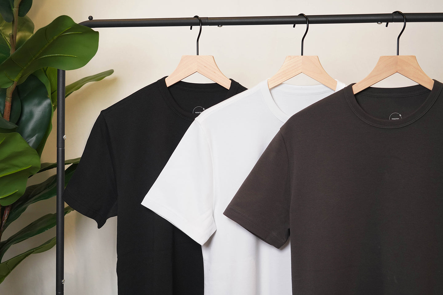 black, white, and charcoal MakTee shirts hanging from a rack next to a plant